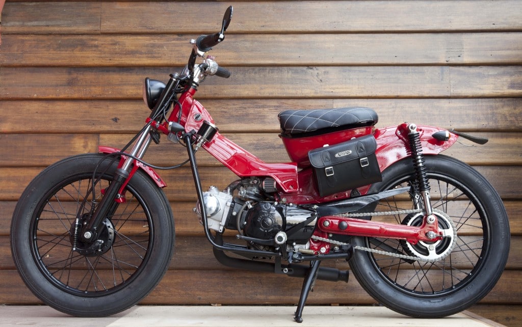 A custom Honda CT110 'Postie' bike against a wood panel background